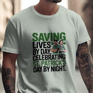 May include: A light green t-shirt with the text "SAVING LIVES BY DAY, CELEBRATING ST. PATRICK'S DAY BY NIGHT." The shirt features a graphic of two medical professionals and shamrocks. The shirt is being worn.