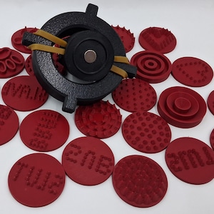 May include: A black metal tool with a circular design and a magnet, secured with a rubber band. Surrounding the tool are numerous red circular stamps with various patterns and text, ideal for crafting or design projects.