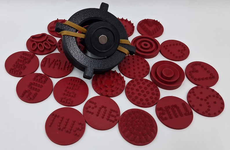 May include: A black metal tool with a circular design and a magnet, secured with a rubber band. Surrounding the tool are numerous red circular stamps with various patterns and text, ideal for crafting or design projects.