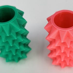 May include: Two cylindrical objects with a textured, spiky exterior surface. One object is a solid green color, and the other is a solid coral pink color. Both items feature a hollow, circular opening at the top and a repeating geometric pattern.
