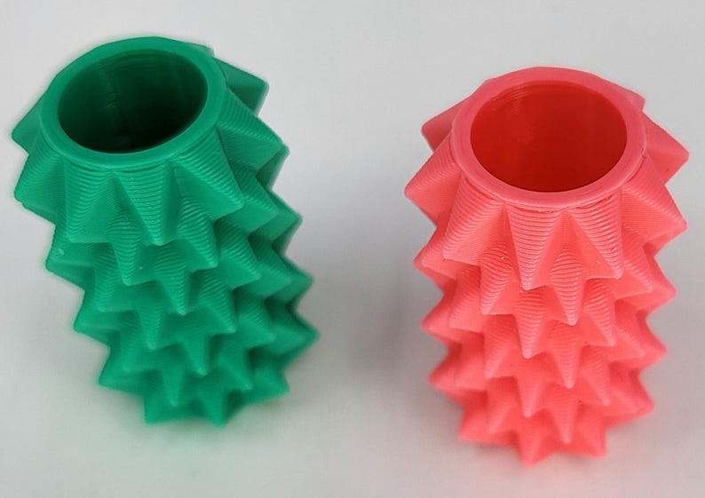 May include: Two cylindrical objects with a textured, spiky exterior surface. One object is a solid green color, and the other is a solid coral pink color. Both items feature a hollow, circular opening at the top and a repeating geometric pattern.
