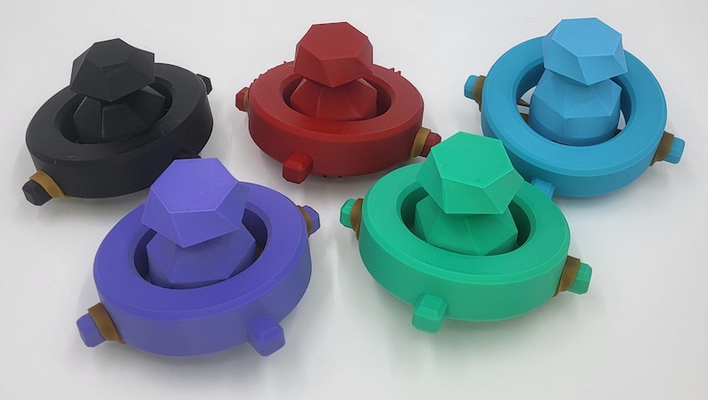 May include: Five colorful, geometric bottle stoppers. Each stopper features a central, faceted gem shape atop a circular base. Colors include black, red, blue, purple, and green. The stoppers have small, cylindrical accents.