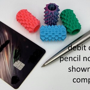 May include: Four cylindrical pencil grips with textured, spiked surfaces in coral, purple, green, and blue. A black debit card and a silver mechanical pencil are positioned nearby for size comparison, with text stating debit card and pencil not included.