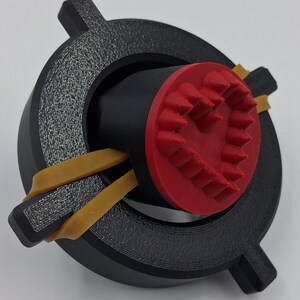 May include: A 3D-printed device featuring a black circular frame with textured detailing and two protruding tabs. Inside, a black cylinder is capped with a red, heart-shaped design. Yellow rubber bands secure the components.