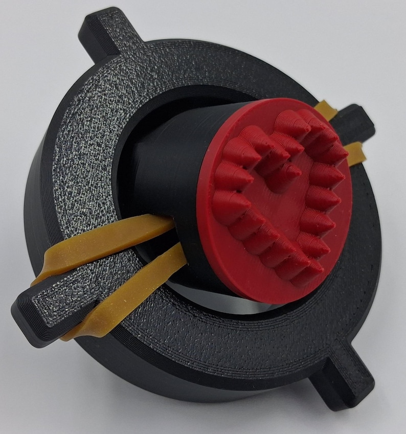 May include: A 3D-printed device featuring a black circular frame with textured detailing and two protruding tabs. Inside, a black cylinder is capped with a red, heart-shaped design. Yellow rubber bands secure the components.