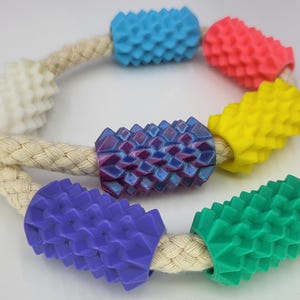 May include: A braided beige rope threaded with seven cylindrical beads featuring a textured, spiky surface. The beads are colored in white, light blue, pink, yellow, purple, dark purple, and green, arranged along the length of the rope.