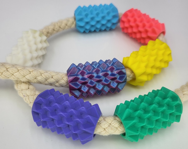 May include: A braided beige rope threaded with seven cylindrical beads featuring a textured, spiky surface. The beads are colored in white, light blue, pink, yellow, purple, dark purple, and green, arranged along the length of the rope.
