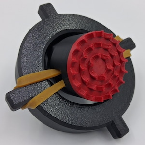 May include: A 3D-printed object featuring a black circular frame with textured surface and a red, gear-shaped center. Two tan rubber bands are secured around the frame. The object is designed with a focus on mechanical components and geometric shapes.