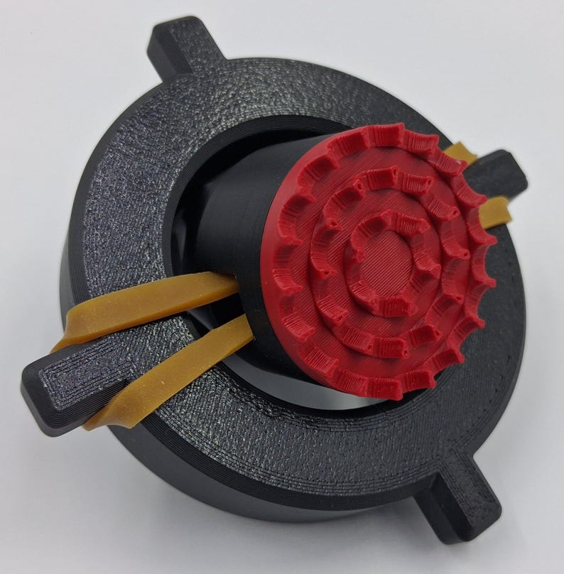 May include: A 3D-printed object featuring a black circular frame with textured surface and a red, gear-shaped center. Two tan rubber bands are secured around the frame. The object is designed with a focus on mechanical components and geometric shapes.