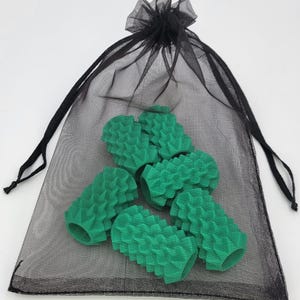 May include: A black sheer drawstring mesh bag containing five cylindrical green pencil grips. Each grip features a textured, spiked surface pattern and a hollow center, designed to slide onto a standard writing instrument for added grip.