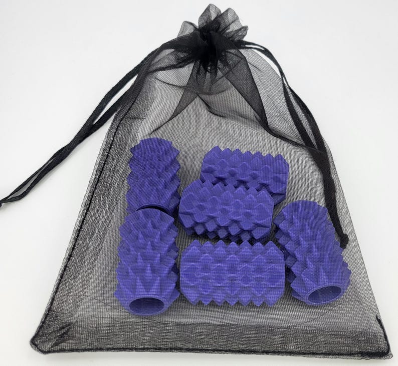 May include: A set of five purple cylindrical objects with a textured, spiked surface, contained within a sheer black drawstring mesh bag. Each purple piece features a hollow center and a repeating geometric pattern of pointed protrusions.