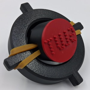 May include: A black, circular object with a red, circular center piece. The red piece has the word "TUBE" embossed on it. Two yellow rubber bands are wrapped around the black object. The object has a textured surface.