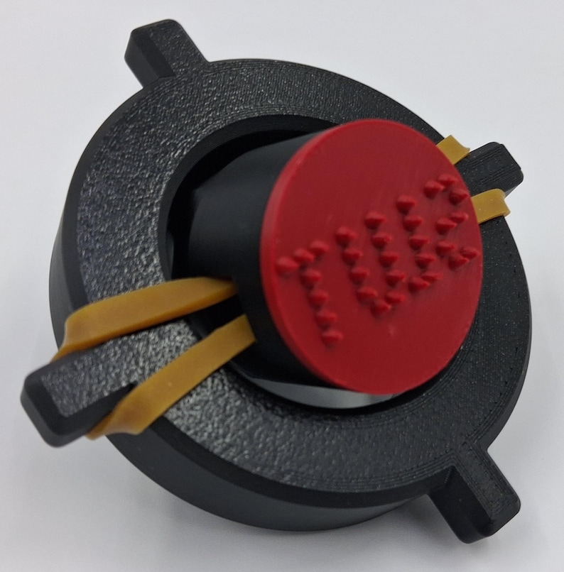 May include: A black, circular object with a red, circular center piece. The red piece has the word "TUBE" embossed on it. Two yellow rubber bands are wrapped around the black object. The object has a textured surface.