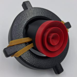 May include: A 3D-printed object featuring a black, circular base with four protruding tabs. A black cylinder is centered within, accented by a red, concentric-ringed element. Two tan rubber bands are stretched across the object, securing the components.