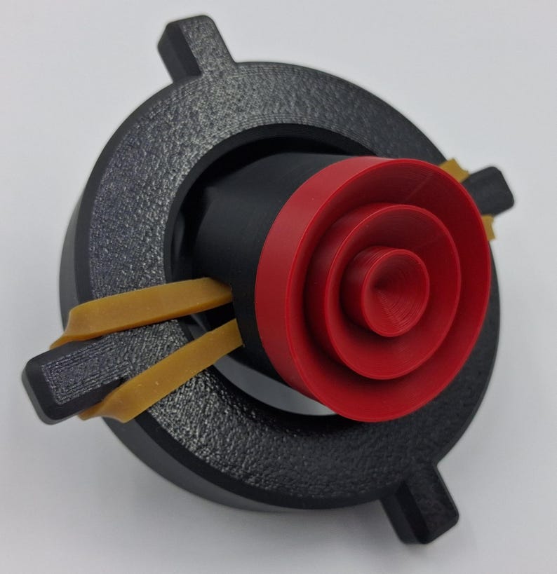 May include: A 3D-printed object featuring a black, circular base with four protruding tabs. A black cylinder is centered within, accented by a red, concentric-ringed element. Two tan rubber bands are stretched across the object, securing the components.