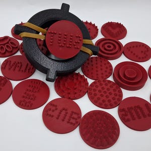 May include: A collection of red, circular stamps with various textured patterns and text, alongside a black, circular stamp holder secured with a rubber band. The stamps are arranged on a white surface.