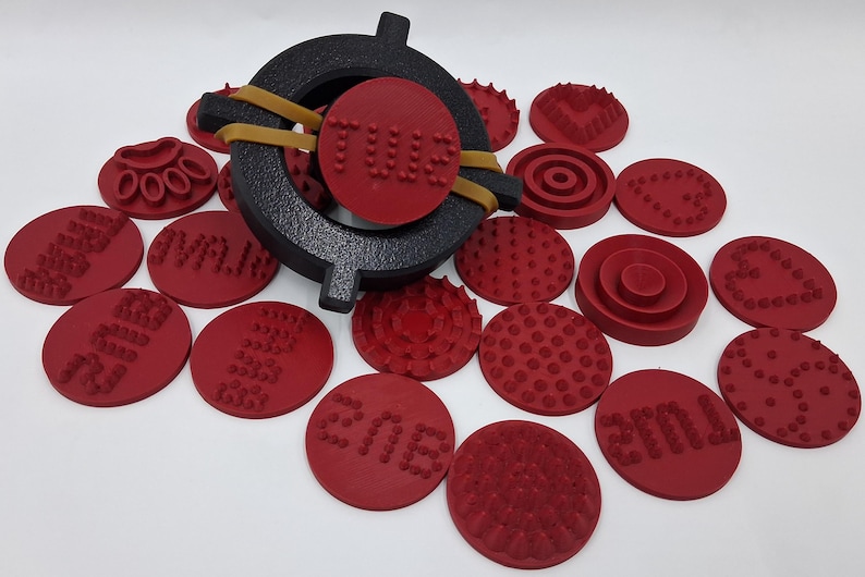 May include: A collection of red, circular stamps with various textured patterns and text, alongside a black, circular stamp holder secured with a rubber band. The stamps are arranged on a white surface.
