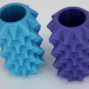 May include: Two cylindrical objects with a textured, spiky, geometric surface pattern. One object is light blue and the other is deep purple. Both items feature a hollow, circular opening at the top and a repeating, layered, ridged design.