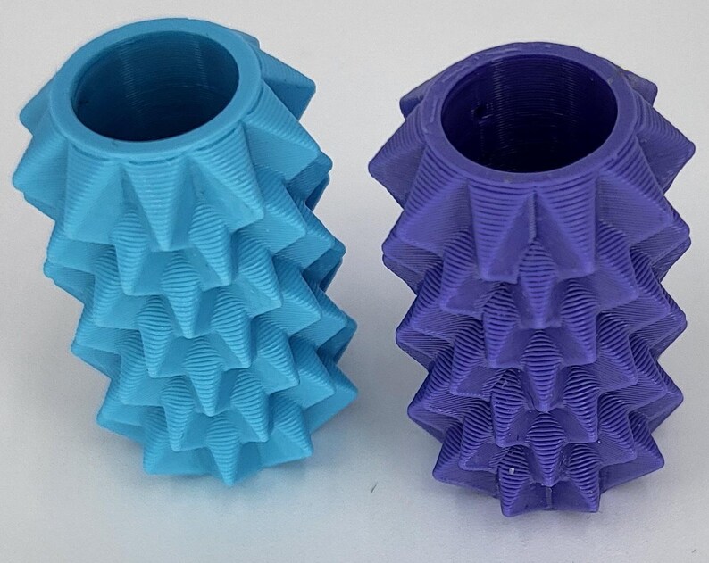 May include: Two cylindrical objects with a textured, spiky, geometric surface pattern. One object is light blue and the other is deep purple. Both items feature a hollow, circular opening at the top and a repeating, layered, ridged design.