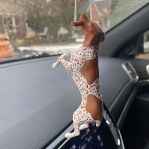 May include: A brown and white dog-shaped car air freshener hanging from a white string. The dog is standing on its hind legs with a spotted pattern on its body. The background shows a car interior and a blurred outdoor scene.
