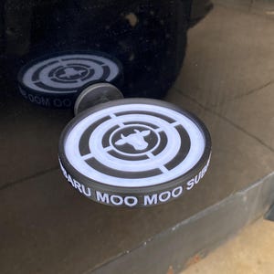 以下が含まれることがあります： 「Subaru Moo Moo Sub」の文字と牛のシルエットが入った、白と黒の丸い車のエンブレム。エンブレムは黒い表面に取り付けられており、反射が見えます。