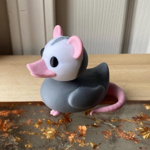 May include: A quirky 3D-printed figurine of an opossum-duck hybrid. The figure has a grey body, a white head with pink ears, a pink beak, and a pink tail. The design is a playful combination of a duck and an opossum.