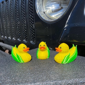 Op de afbeelding: Drie gele rubberen eendjes in de vorm van maïs met groene bladeren. De eendjes zitten op een zwart oppervlak.
