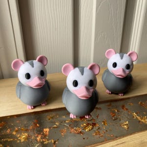 May include: Three grey and white opossum figurines with pink ears and beaks. The figurines are on a wooden surface with gold leaf accents. The opossums have black eyes and are in a row.