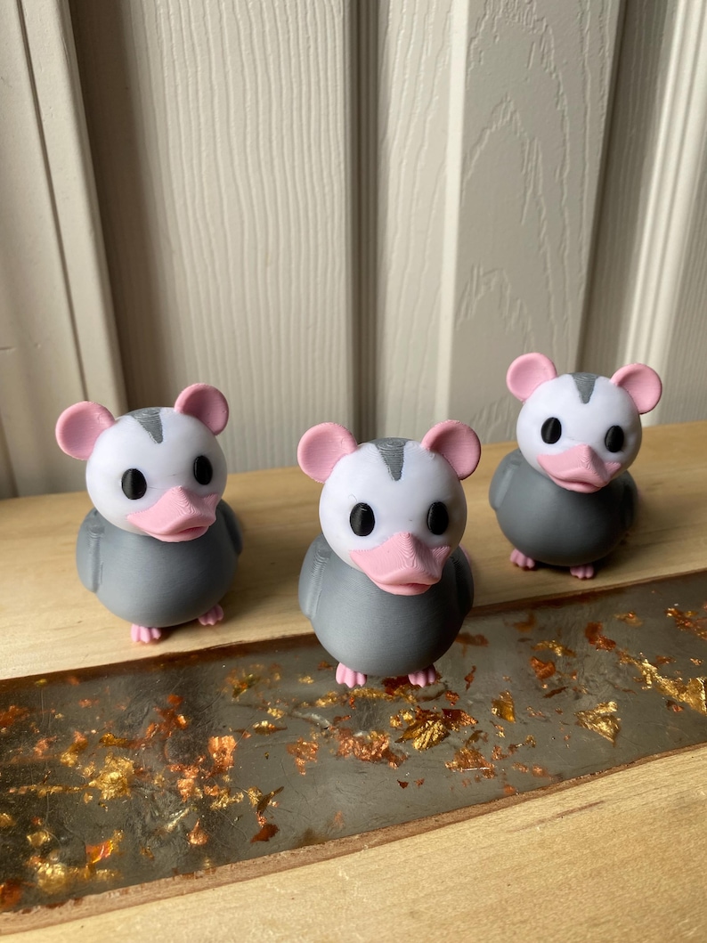 May include: Three grey and white opossum figurines with pink ears and beaks. The figurines are on a wooden surface with gold leaf accents. The opossums have black eyes and are in a row.