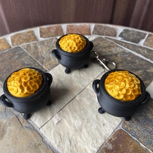 Peut inclure: Trois récipients en forme de chaudron noirs avec des couvercles en forme de pièces d'or. Les récipients sont sur une surface carrelée brune et beige.