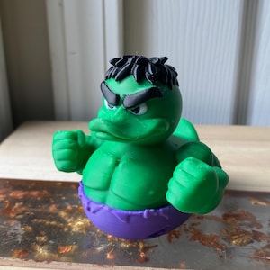 May include: A green rubber duck toy styled as the Hulk, with black hair and eyebrows, and a purple waist. The duck is posed with clenched fists, showcasing its muscular physique. A fun, novelty item.