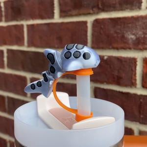 以下が含まれることがあります： 白いストローが上部に挿入された、漫画のクジラの形をしたライトブルーとブラックのシリコン製ストローのトッピング。ストローのトッピングは、ブラウンのハンドルが付いたホワイトとシルバーのトラベルマグの上に置かれています。