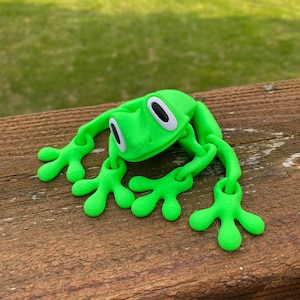 May include: A bright green, 3D-printed frog toy with large, white-rimmed eyes and webbed feet. The frog is lying on a wooden surface, with its limbs outstretched. The toy is a vibrant shade of green.