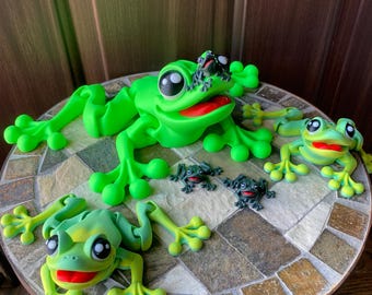 Op de afbeelding: Een set van vijf groene kikkerbeeldjes, in verschillende maten. De grootste kikker ligt met zijn poten gestrekt. De andere kikkers zijn kleiner en zitten of staan op de tafel. De kikkers zijn allemaal gemaakt van een glad, groen materiaal en hebben zwarte ogen en rode monden.