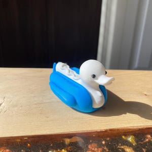 May include: A whimsical 3D-printed duck figurine. The duck has a white head and body, a blue back, and a white, curved shape in the middle. The duck is sitting on a wooden surface.