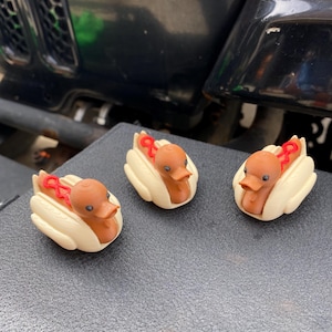 Puede incluir: Tres pequeñas figuras de patos de goma con forma de perritos calientes con mostaza y ketchup. Los patos son marrones con picos blancos y ojos negros. Están sentados sobre una superficie gris.