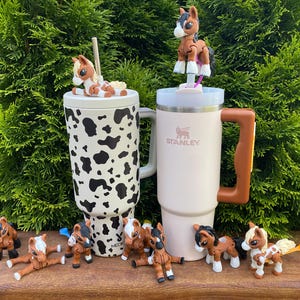 Puede incluir: Dos tazas de viaje aisladas, una con estampado de vaca en blanco y negro y otra en color beige claro, se exhiben con varias figuras de caballos en miniatura. Una figura de caballo descansa sobre cada tapa de la taza. Las tazas tienen asas.