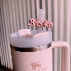 May include: A pink Stanley travel mug with a winged bear logo and the word "STANLEY" printed on the side. The mug has a white lid and a pink handle. A pink straw with a personalised name tag that says "Brooke" is inserted into the mug.