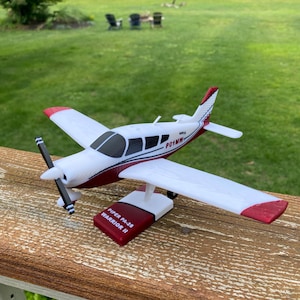 May include: A white and maroon model airplane with a black propeller, featuring the text PO1MW on the side and mounted on a stand labeled PIPER PA-28 WARRIOR II. The aircraft has red wingtips and a red tail section with a white and blue stripe.