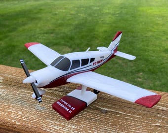 May include: A white and maroon model airplane with a black propeller, featuring the text PO1MW on the side and mounted on a stand labeled PIPER PA-28 WARRIOR II. The aircraft has red wingtips and a red tail section with a white and blue stripe.