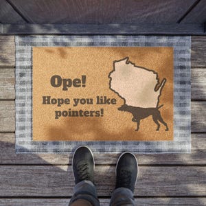 May include: A rectangular doormat with a tan centre and a grey plaid border. The text Ope! Hope you like pointers! is printed in dark brown. A dark brown silhouette of the state of Wisconsin and a pointer dog are featured on the right side.