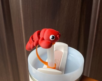 May include: A white Stanley travel mug with a brown handle and a red lobster-shaped straw topper. The mug has the Stanley logo on the side.