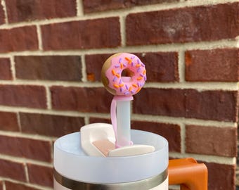 May include: A white Stanley travel mug with an orange handle. The mug has a pink donut-shaped straw topper with purple and orange sprinkles.
