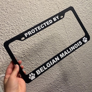 Puede incluir: Marco de matrícula negro con texto blanco que dice "PROTECTED BY" en la parte superior y "BELGIAN MALINOIS" en la parte inferior. Huellas de patas en la parte inferior derecha e izquierda. El marco está sostenido por una mano.