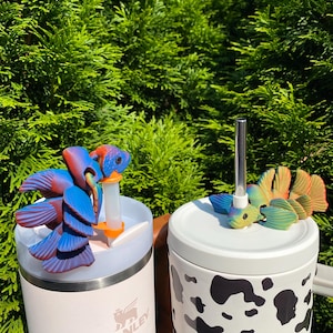 Puede incluir: Dos vasos aislados con adornos en forma de pájaro. Uno es blanco con estampado de vaca negro, el otro es rosa claro con el logotipo de Stanley. Los adornos de pájaros son azules, naranjas y verdes.