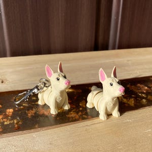May include: Two small, cream-coloured dog figurines with pink ears and noses. One has a silver keychain attached. The dogs are posed on a wooden surface with a metallic strip. The background is a brown wooden panel.