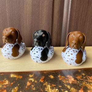 Könnte beinhalten: Drei hundeförmige Figuren. Die Hunde haben braune oder schwarze Köpfe und Ohren mit weiß-braun gefleckten Körpern. Die Figuren stehen auf einer Holzoberfläche vor einem braunen Hintergrund.