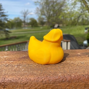 May include: A bright yellow rubber duck toy with a unique design. The duck is positioned on a weathered wooden surface, with a blurred outdoor background of trees and sky. The duck's design is a playful take on the classic rubber duck.