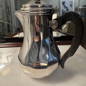 May include: A silver coffee pot with a wooden handle. The pot has a rounded body and a lid with a decorative knob.
