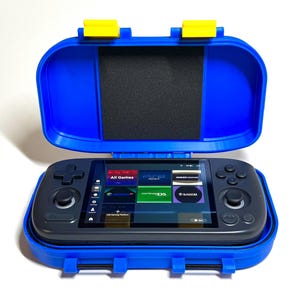 May include: A handheld gaming console with a screen displaying game icons, inside an open blue case with yellow clasps. The console is black with control buttons and a directional pad. The case has a black foam insert.